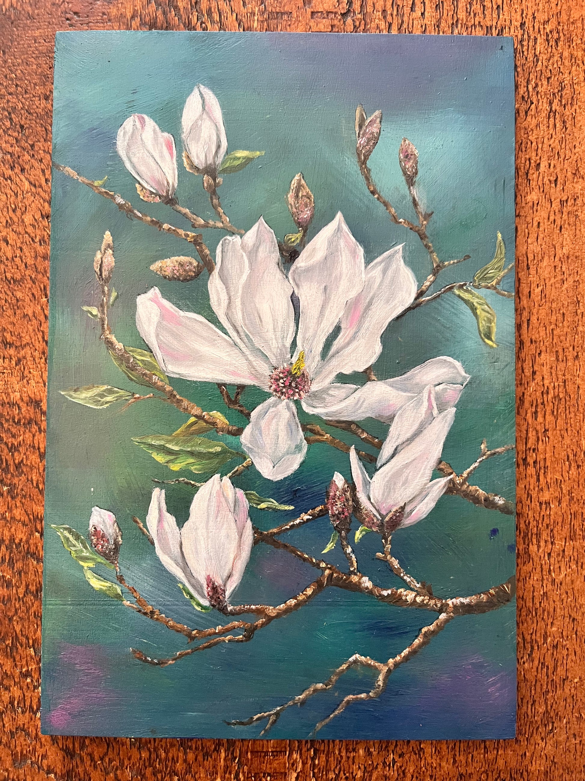 Painting of white flowers on a branch with a teal background, placed on a wooden surface.